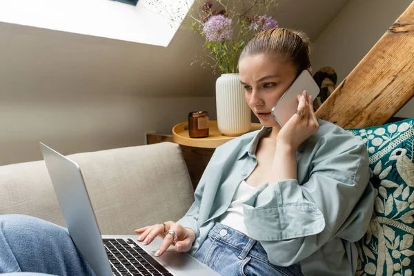 Young woman talking on smartphone and using laptop on couch at home — Stock Photo