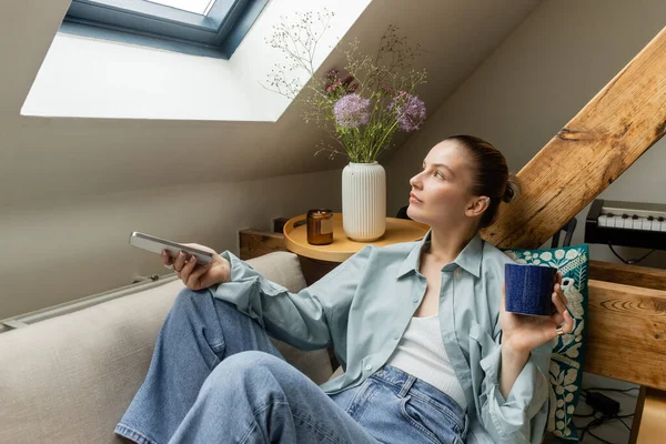 Junge Frau sitzt zu Hause auf Couch und hält Tasse und Smartphone in der Hand — Stockfoto