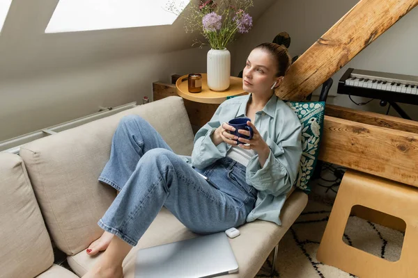 Mujer joven en auricular sosteniendo la taza cerca de la computadora portátil en el sofá en casa - foto de stock