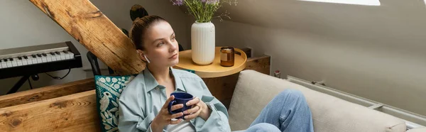 Жінка в навушниках тримає чашку, сидячи на дивані вдома, банер — стокове фото