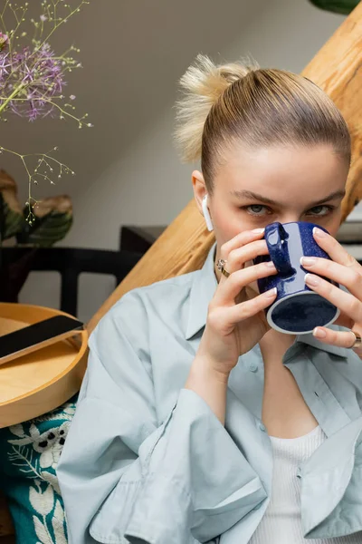 Mujer joven en auriculares bebiendo café cerca de teléfono inteligente con pantalla en blanco en casa - foto de stock