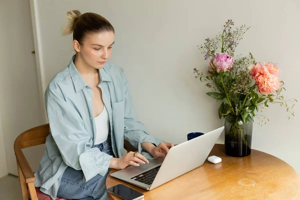 Jovem mulher usando laptop perto de copo e flores em casa — Fotografia de Stock