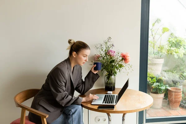 Joven freelancer usando laptop y sosteniendo taza cerca de flores en casa - foto de stock