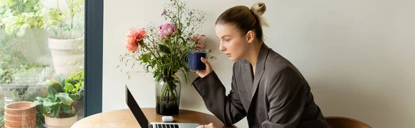 Mujer con chaqueta sosteniendo la taza y el uso de la computadora portátil cerca de las flores en casa, bandera - foto de stock