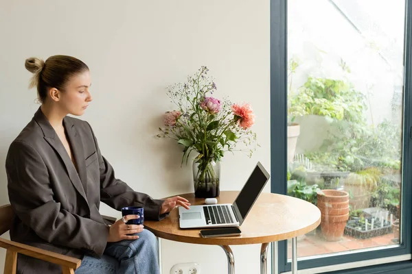 Vista lateral de la mujer en la chaqueta que sostiene la taza y el uso de dispositivo cerca de las flores en casa - foto de stock