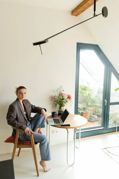 Mujer joven con chaqueta mirando a la cámara cerca de dispositivos y flores en casa - foto de stock