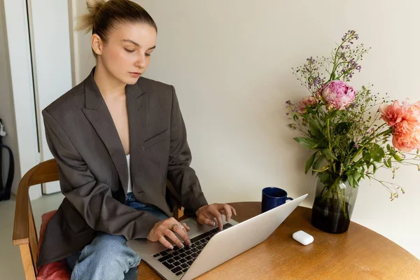 Joven freelancer usando laptop cerca de taza y ramo en casa - foto de stock