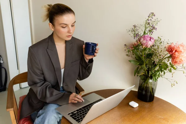 Jeune femme en blazer utilisant un ordinateur portable et tenant la tasse près du bouquet sur la table à la maison — Photo de stock