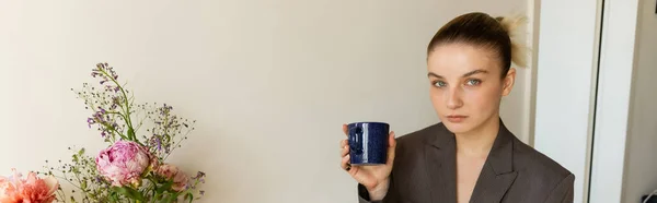 Young woman in blazer holding cup and looking at camera near flowers at home, banner — Stock Photo