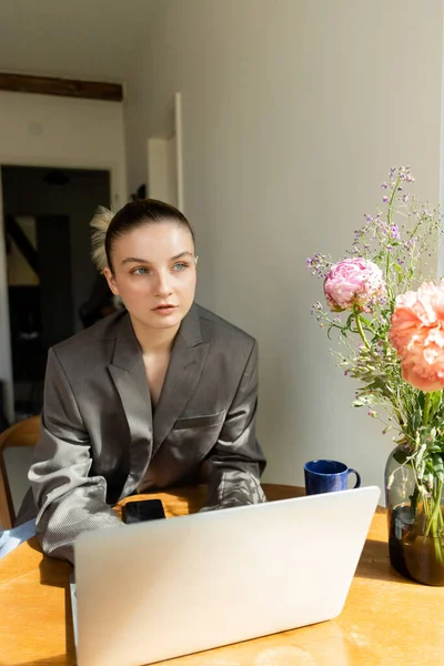 Young woman in blazer looking at flowers near devices and cup at home — Stock Photo