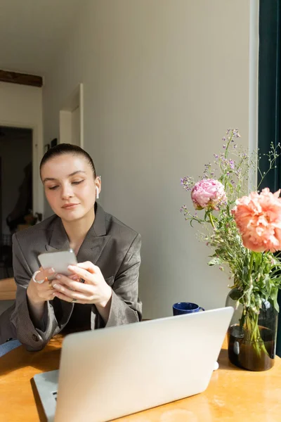 Jeune femme dans l'écouteur en utilisant smartphone près de l'ordinateur portable et bouquet sur la table à la maison — Photo de stock