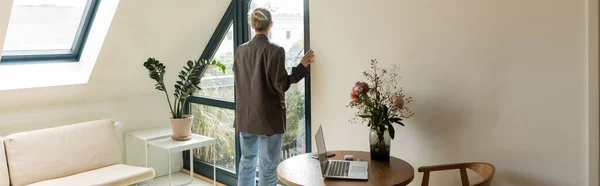 Вид сзади женщины, стоящей возле двери балкона и устройств на столе в домашних условиях, баннер — стоковое фото
