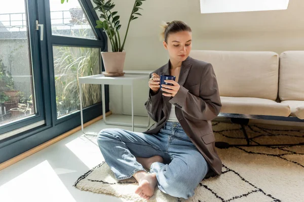 Mujer con chaqueta sosteniendo la taza mientras está sentado en la alfombra en la sala de estar - foto de stock