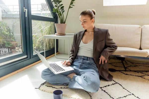 Young freelancer in jacket using laptop near cup on carpet at home — Stock Photo