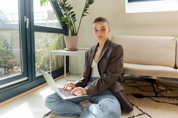 Mujer joven en chaqueta usando el ordenador portátil y mirando a la cámara en la alfombra en casa - foto de stock