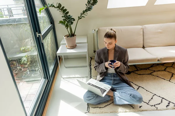 Freelancer em blazer segurando copo e olhando para laptop no tapete em casa — Fotografia de Stock