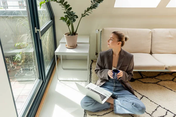 Joven freelancer sosteniendo la taza y mirando las ventanas cerca del portátil en la alfombra en casa - foto de stock