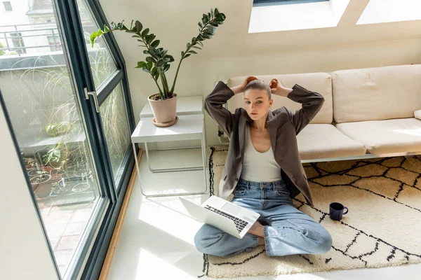 Freelancer in jacket touching hair near laptop and cup on floor at home — Stock Photo