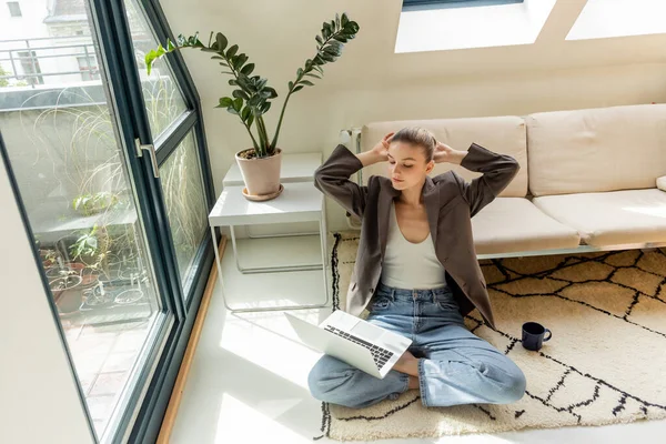 Femme en veste regardant ordinateur portable près de tasse sur le tapis à la maison — Photo de stock