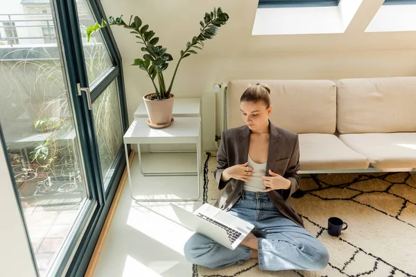 Freelancer en chaqueta mirando portátil cerca de la taza en el sofá en la sala de estar - foto de stock