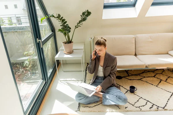 Joven freelancer en chaqueta hablando en smartphone y usando portátil cerca de taza en alfombra en casa - foto de stock