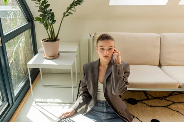 Junge Frau im Blazer spricht mit Smartphone und hält Laptop im Wohnzimmer — Stockfoto