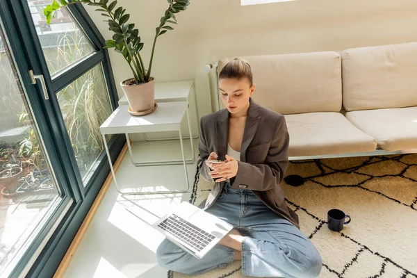 Junge Frau in Jacke und Jeans mit Smartphone und Laptop in der Nähe Tasse auf Teppich zu Hause — Stockfoto
