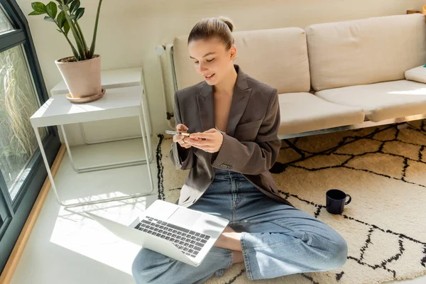 Freelance sorridente che utilizza laptop e smartphone vicino a tazza su tappeto su moquette — Foto stock