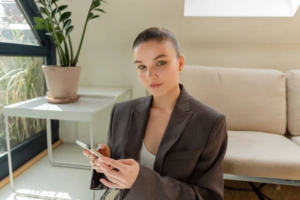 Mujer joven con chaqueta sosteniendo el teléfono inteligente y mirando a la cámara en la sala de estar - foto de stock