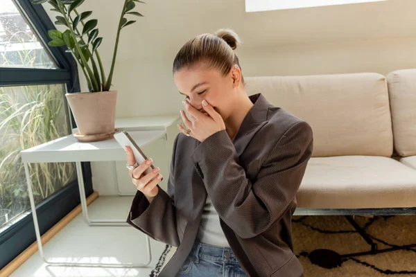 Fröhliche Frau in Jacke mit Smartphone im Wohnzimmer — Stockfoto