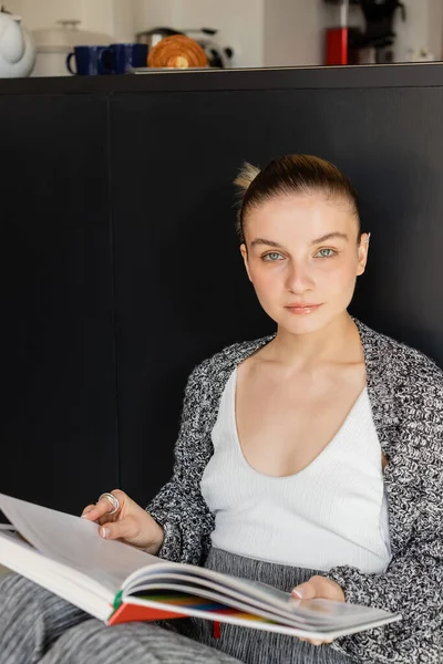 Junge Frau hält Buch in der Hand und blickt zu Hause in die Kamera — Stockfoto