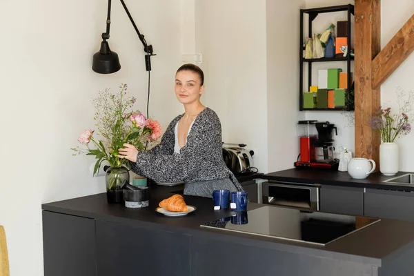 Mujer sonriente poniendo flores en jarrón cerca de copas y croissant en la cocina - foto de stock