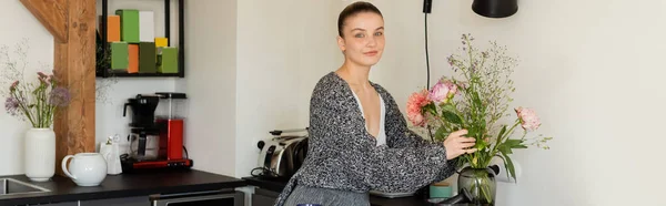 Жінка в в'язаному кардигані кладе букет у вазу на кухні, банер — стокове фото