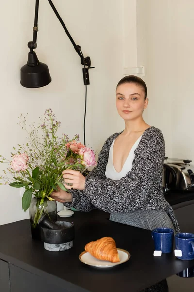 Donna in cardigan lavorato a maglia che mette fiori in vaso vicino a croissant e tè in cucina — Foto stock