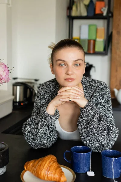 Young woman in knitted cardigan looking at camera near croissant and cups in kitchen — Stock Photo