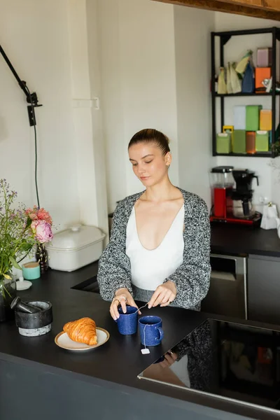 Mulher em cardigan segurando xícara com saco de chá perto de croissant na cozinha — Fotografia de Stock
