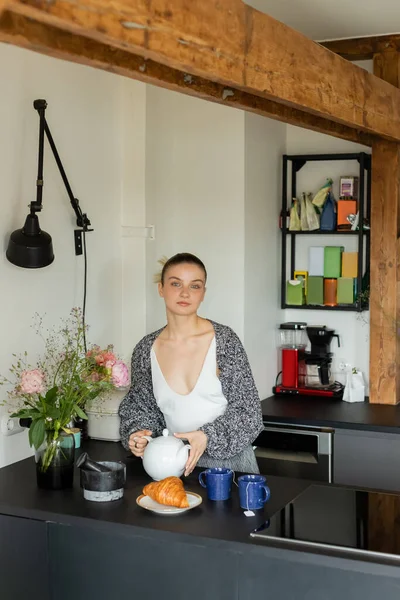 Young woman in knitted cardigan holding teapot near cups and croissant in kitchen — Stock Photo