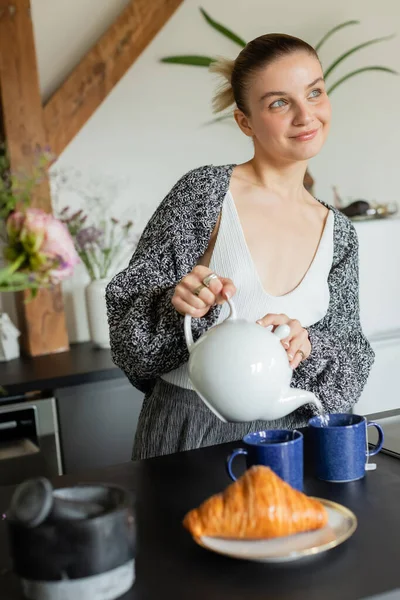 Donna positiva in maglia cardigan versando tè vicino a croissant in cucina — Foto stock