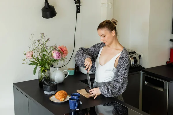 Donna in maglia cardigan taglio avocado vicino croissant e tazze in cucina — Foto stock