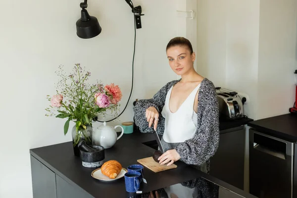 Young woman in warm cardigan cutting avocado near tea and croissant in kitchen — Stock Photo