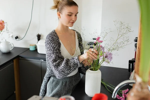 Jovem mulher em cardigan de malha colocando flores em vaso em casa — Fotografia de Stock