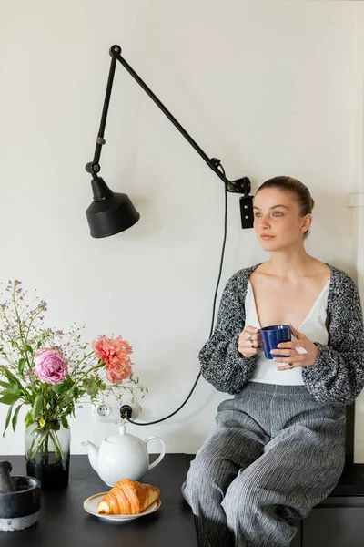 Young woman in warm cardigan holding cup near croissant and teapot in kitchen — Stock Photo