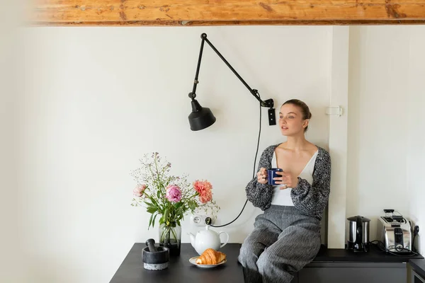 Donna in maglia cardigan tenendo tazza vicino croissant e fiori in cucina — Foto stock