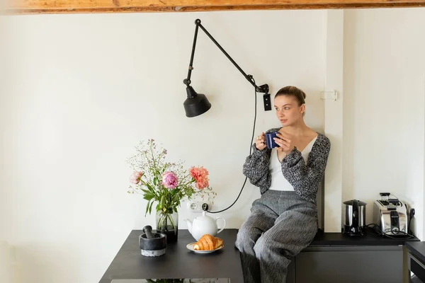 Donna in caldo cardigan in possesso di tazza vicino a croissant e fiori in cucina a casa — Foto stock