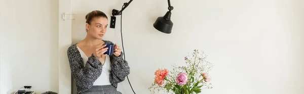 Donna in cardigan che tiene una tazza di tè vicino a lampada e fiori a casa, striscione — Foto stock
