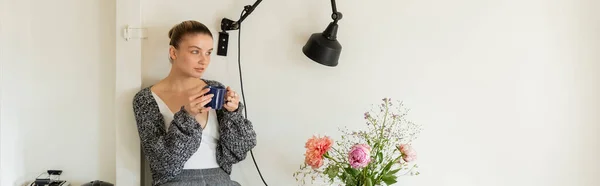 Mujer en cárdigan sosteniendo taza de té cerca de la lámpara y flores en casa, pancarta - foto de stock