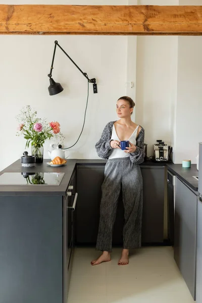 Junge Frau in Strickjacke hält Tasse in moderner Küche zu Hause — Stockfoto