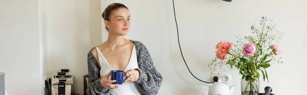 Mujer sosteniendo la taza cerca de la tetera y flores en la cocina moderna, pancarta — Stock Photo