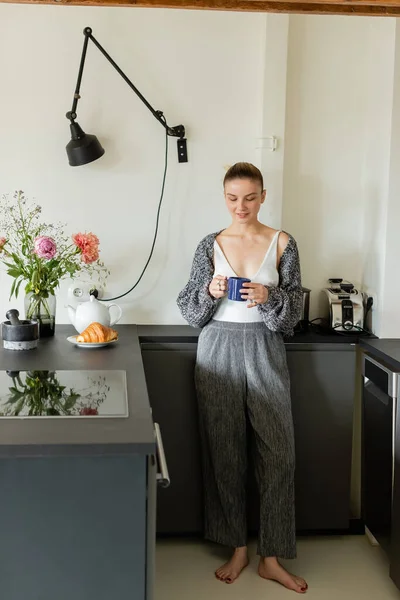 Mujer sonriente en un cálido cárdigan sosteniendo taza cerca de croissant y tetera en la cocina — Stock Photo