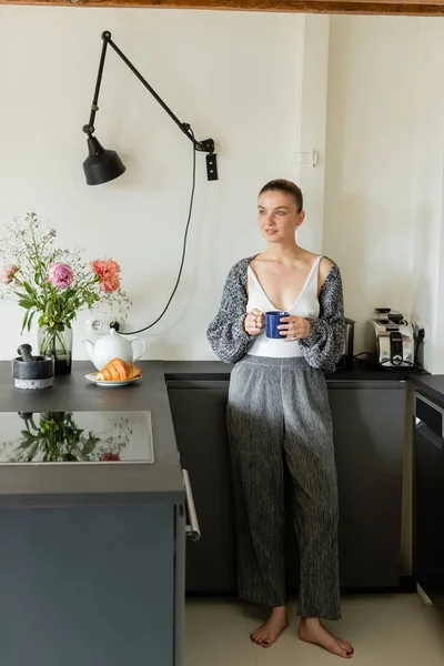 Positive woman holding cup and looking away near croissant in modern kitchen — Stock Photo